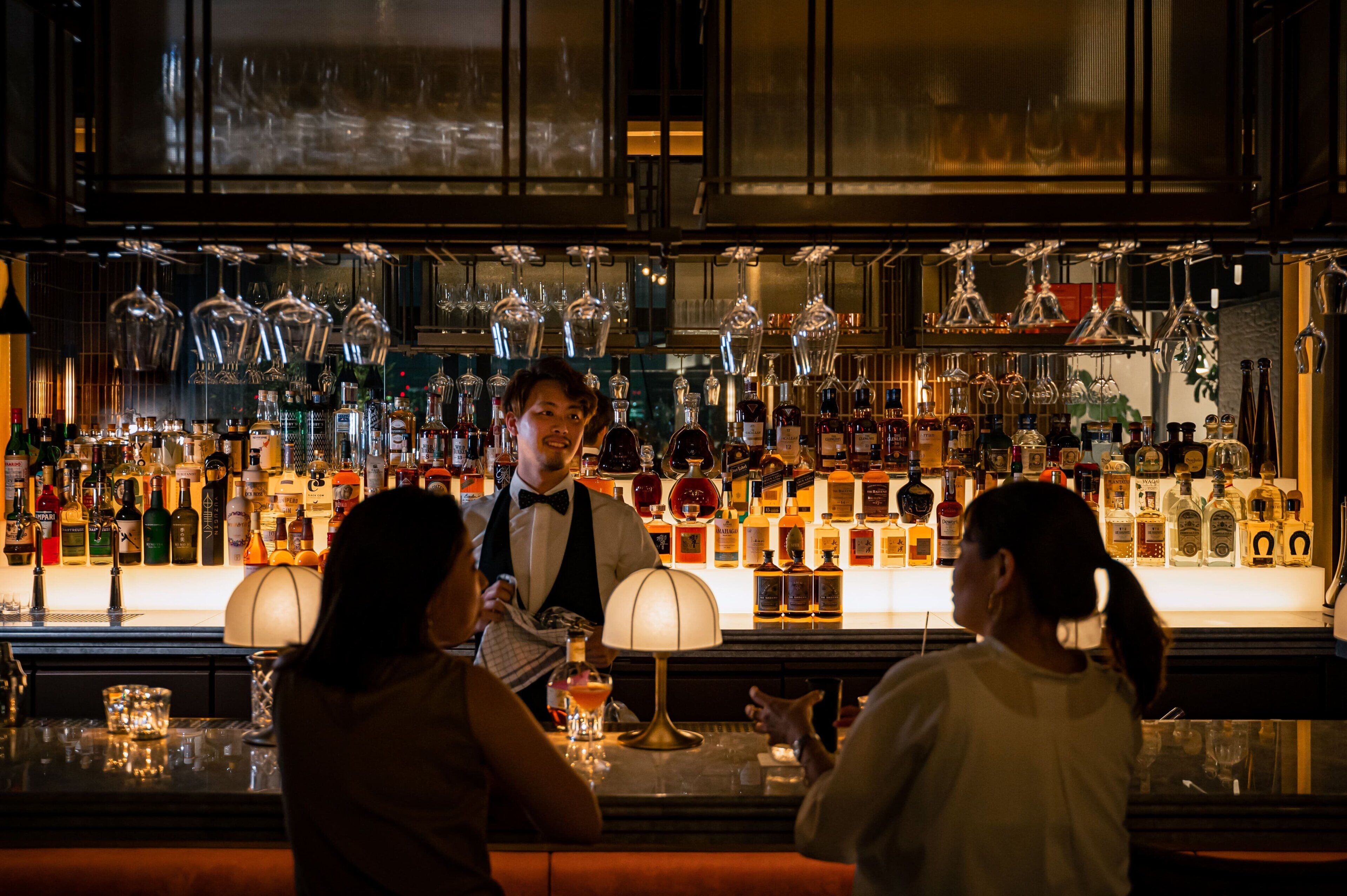 A bar with a bartender and two customers. The bar has a variety of liquor bottles and glassware.