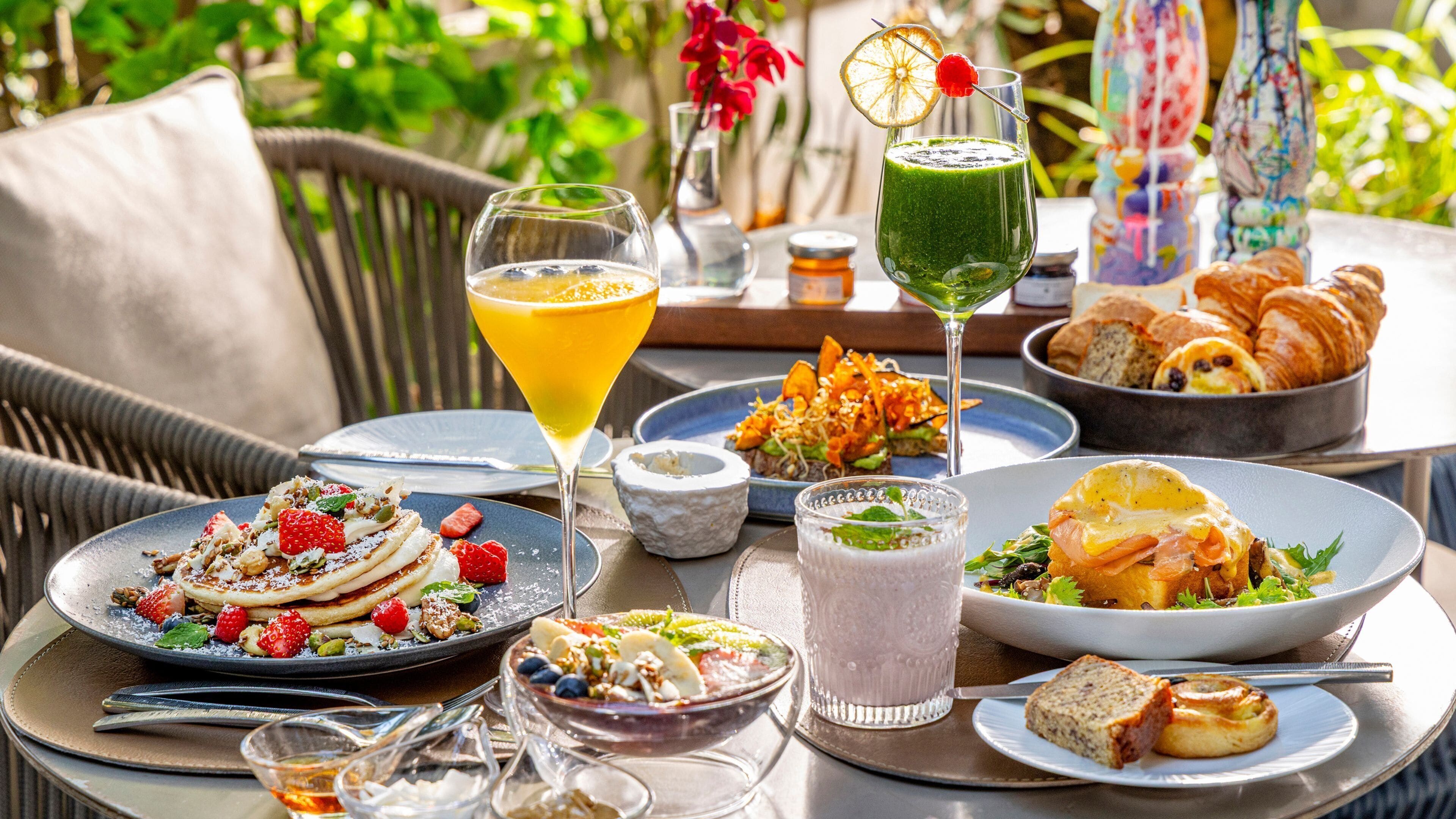 A table set with various dishes, drinks, and pastries, likely in an outdoor dining area with greenery in the background.