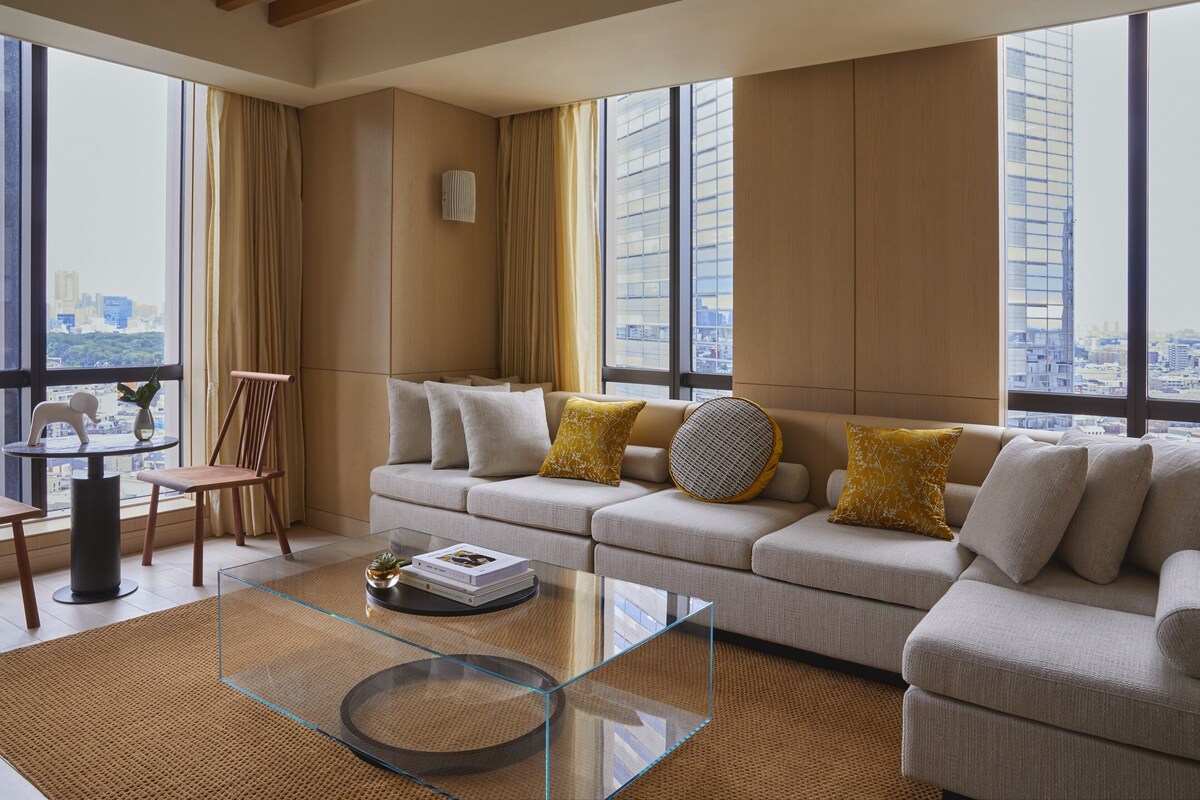 Una sala de estar moderna con un sofá grande, una mesa de centro de vidrio y vistas a un paisaje urbano a través de amplios ventanales.