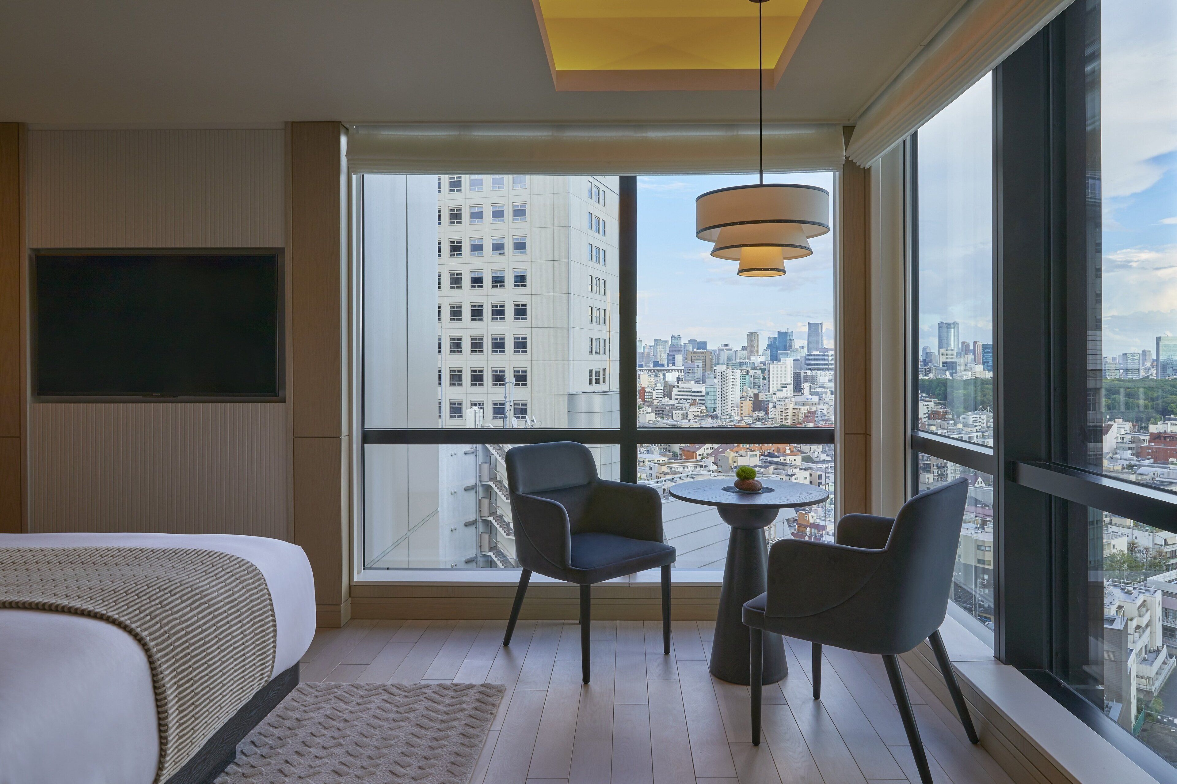 Habitación de hotel moderna con un ventanal grande que ofrece vistas a la ciudad, una cama, una mesita con sillas y un televisor montado en la pared.