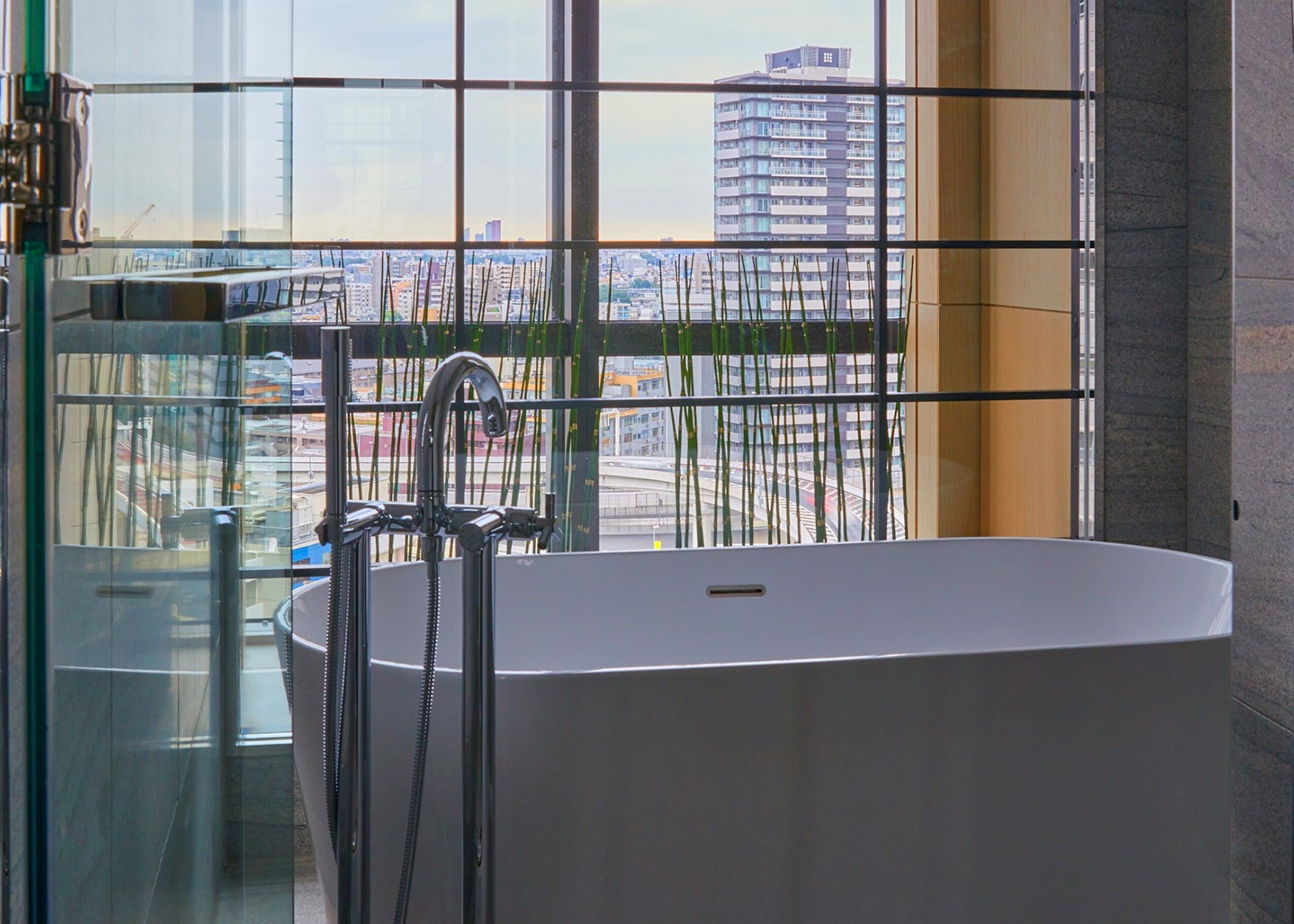 A modern bathroom with a freestanding bathtub, a glass shower enclosure, and a view of a cityscape through large windows.