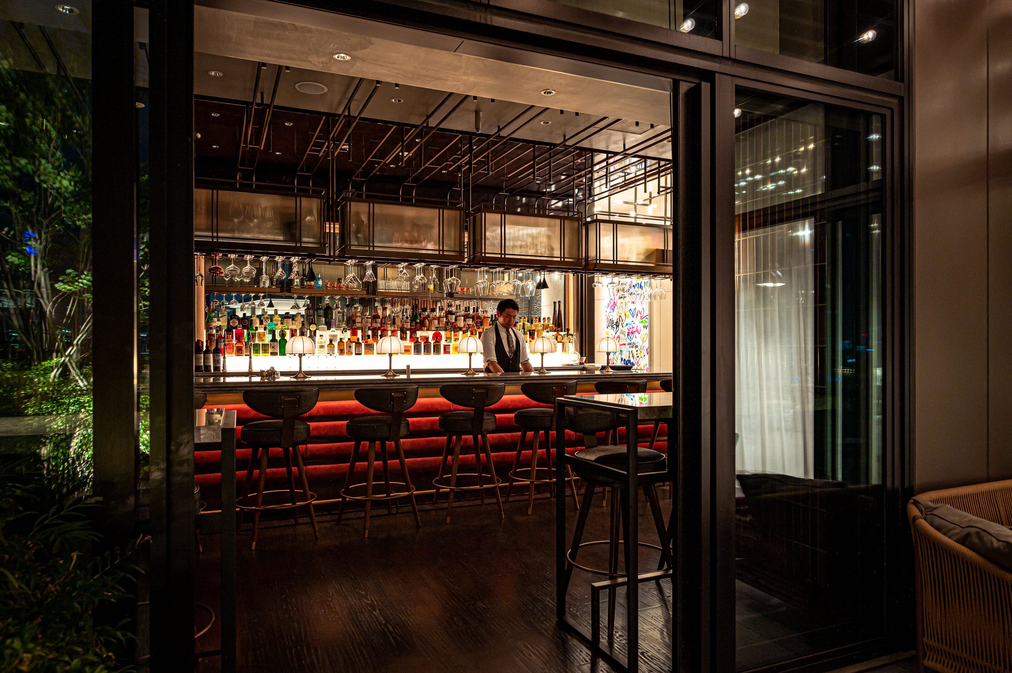 An interior bar area with a bartender, red bar stools, and a variety of bottles and glasses.