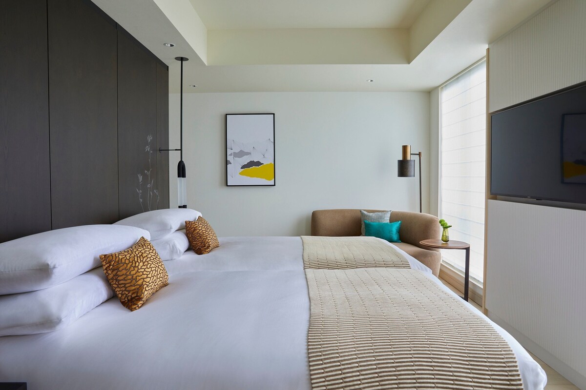 Una habitación de hotel moderna con una cama grande, un sofá, un televisor y una ventana con persianas.