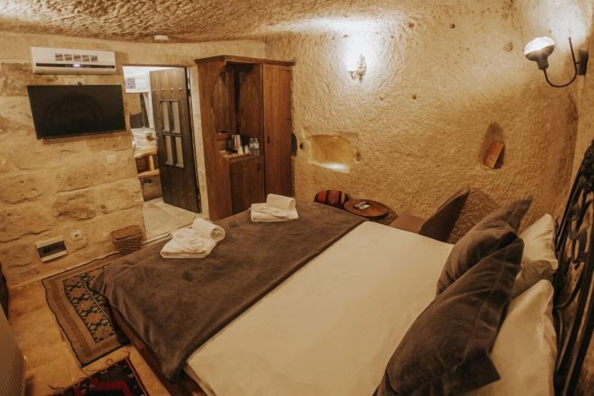 A stone-walled room with a bed, a wooden wardrobe, a television, and a wall-mounted light fixture.