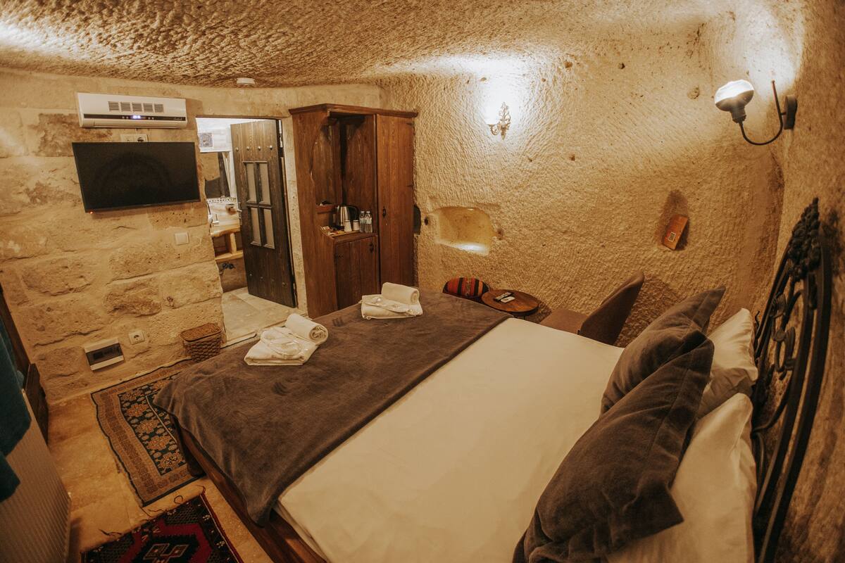 A room with a bed, a flat-screen TV mounted on the wall, a wooden cabinet, and a small table with a lamp.