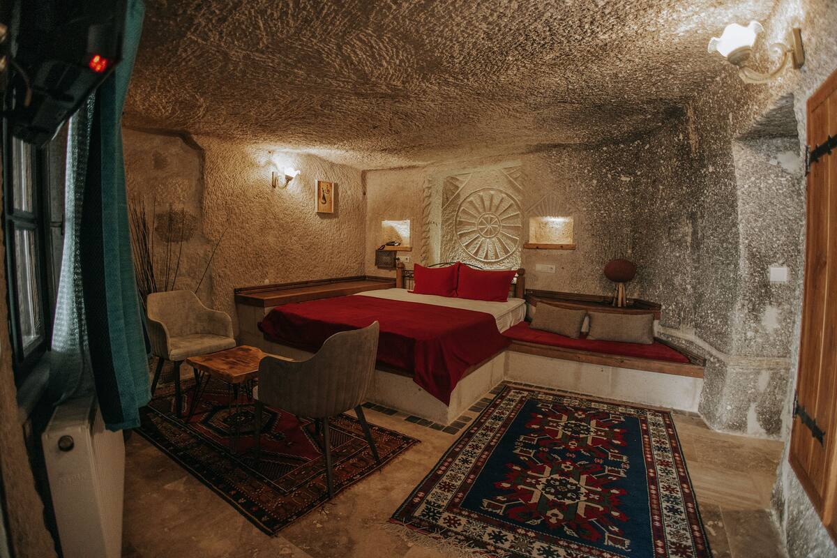 A rustic stone room with a bed, a chair, a small table, and a rug.
