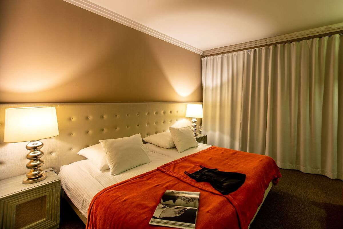 A hotel room with a large bed, two bedside lamps, a nightstand, a magazine on the bed, and a window with curtains.