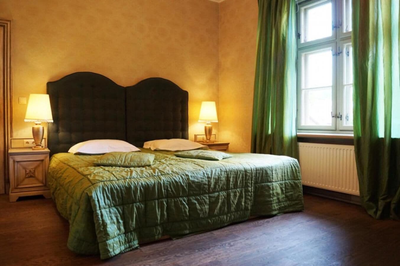 A bedroom with a large bed, two bedside tables with lamps, a radiator, and green curtains.