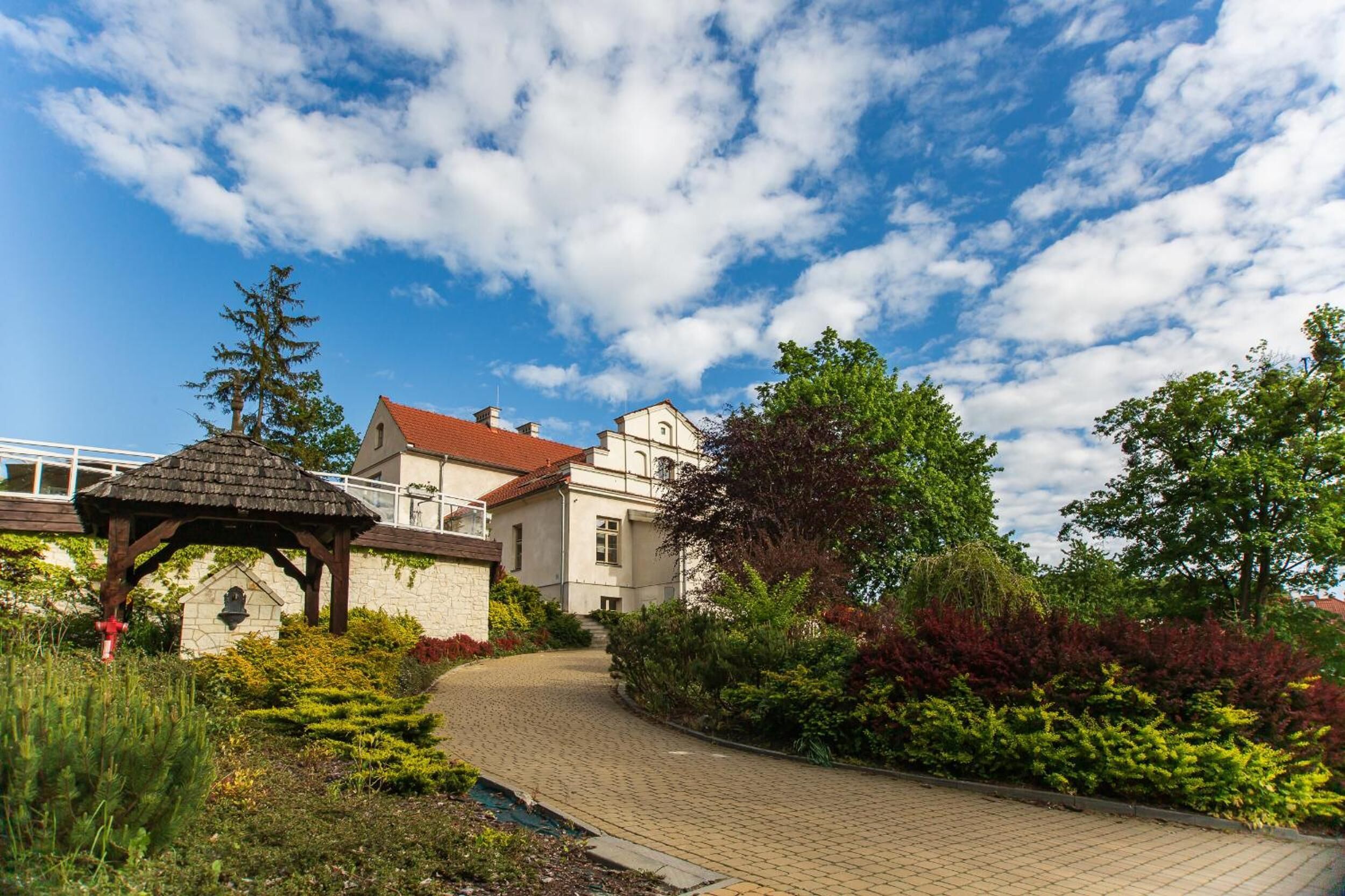 A garden pathway leading to a building with a red roof, surrounded by lush greenery and a gazebo.