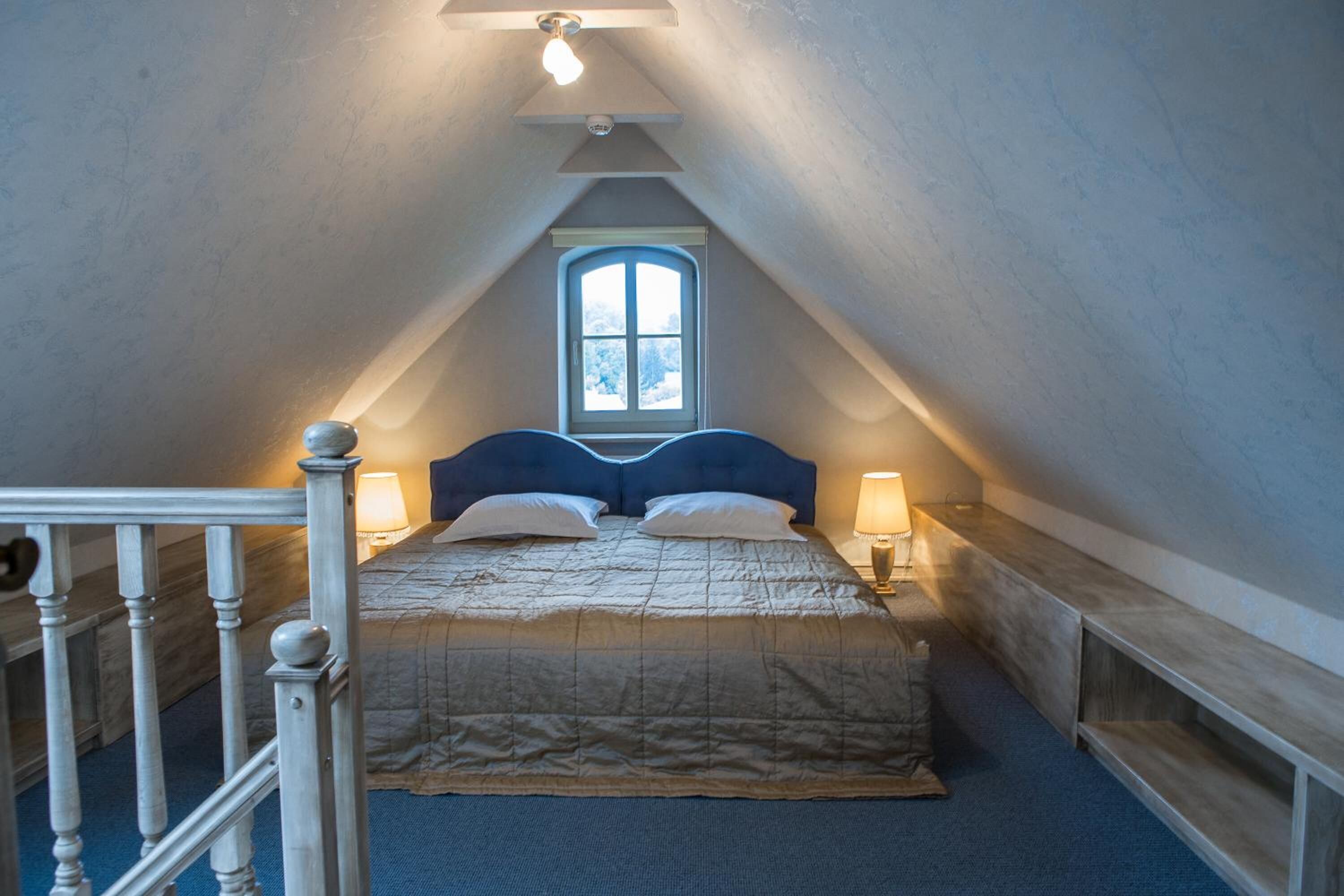 Ein Schlafzimmer mit schrägem Dach, einem Fenster, einem Bett mit Kopfteil, Nachttischlampen und einer hölzernen Bank.