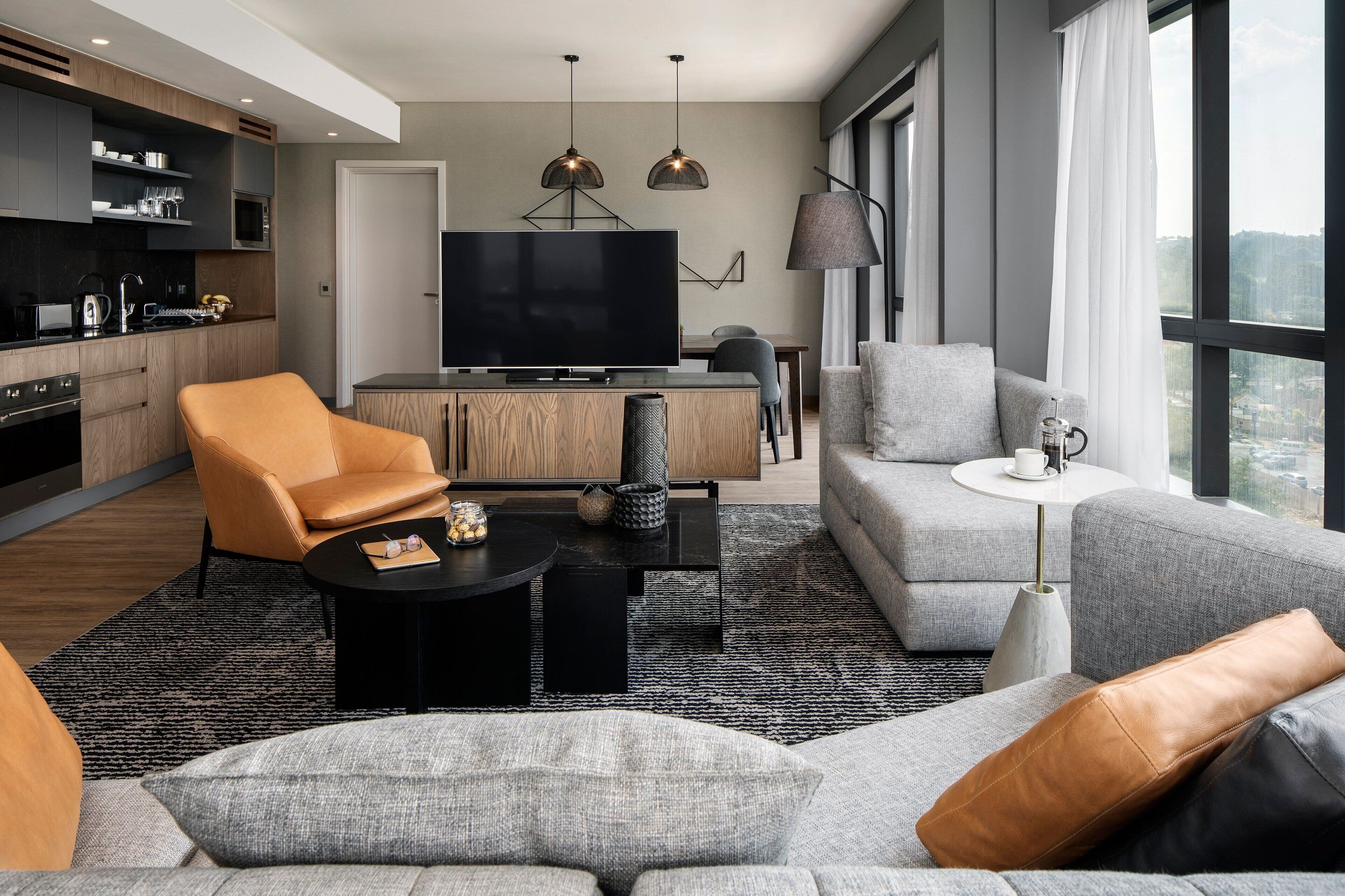 Una sala moderna con sofá, sillón y mesa de centro. Se observa televisión y un comedor al fondo.