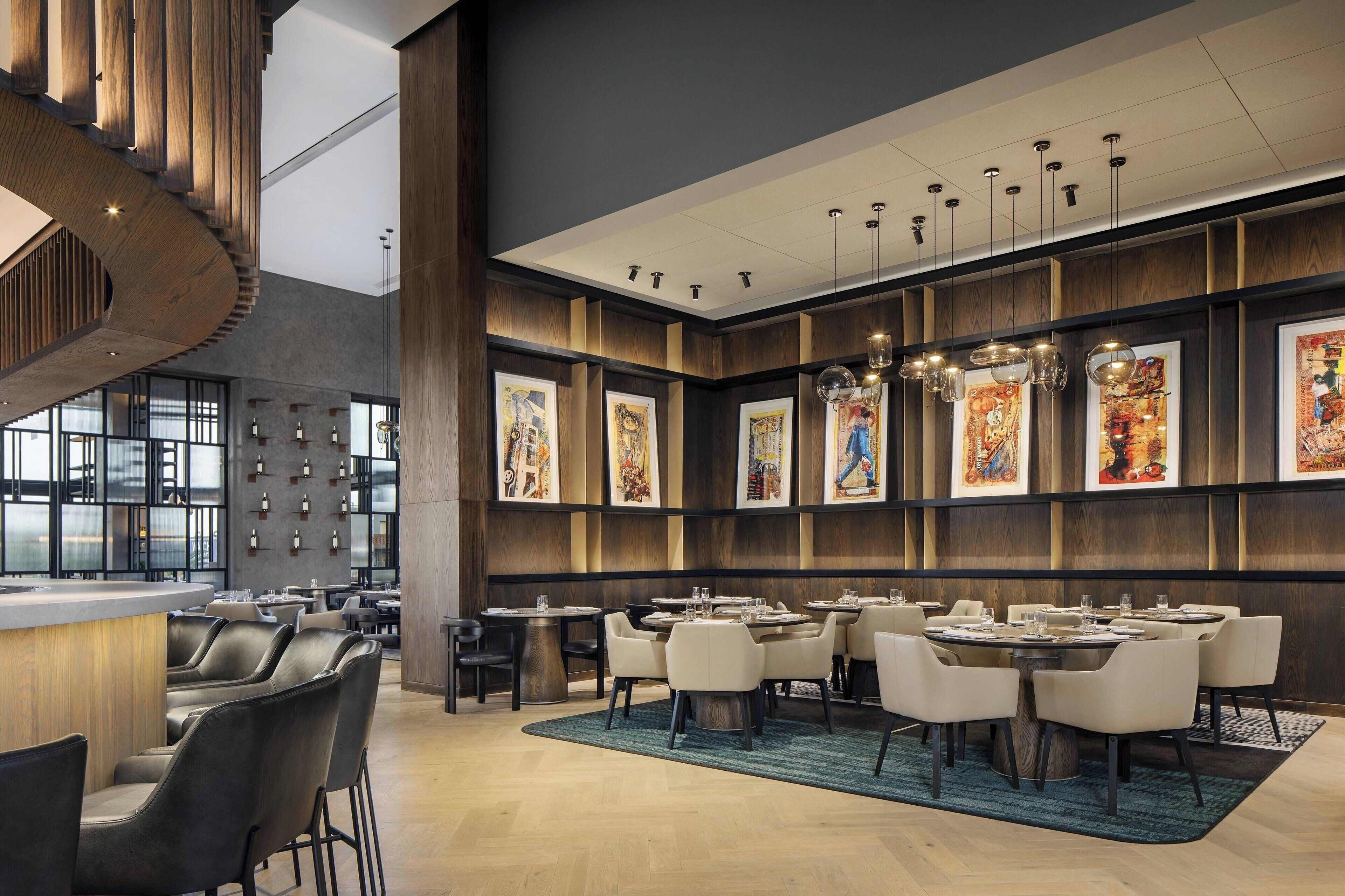 Un restaurant moderne avec de hauts plafonds, des boiseries et des œuvres d’art accrochées aux murs. Les tables sont disposées pour accueillir les clients, et un bar avec des tabourets est également à leur disposition.