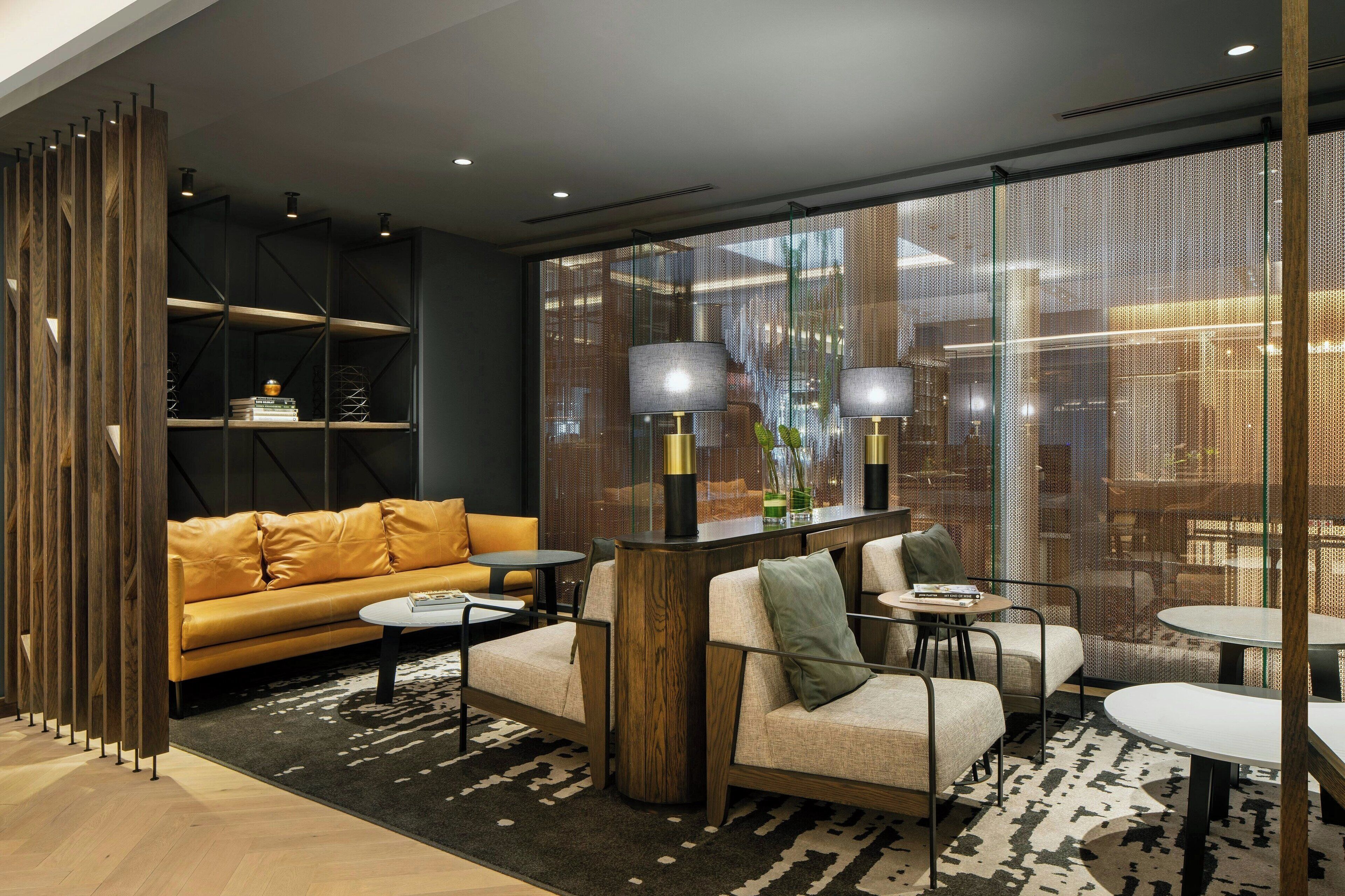 A modern living room with a sofa, armchairs, a coffee table, and a side table. There is a bookshelf with books and decorative items, and a large window with a view of the city.