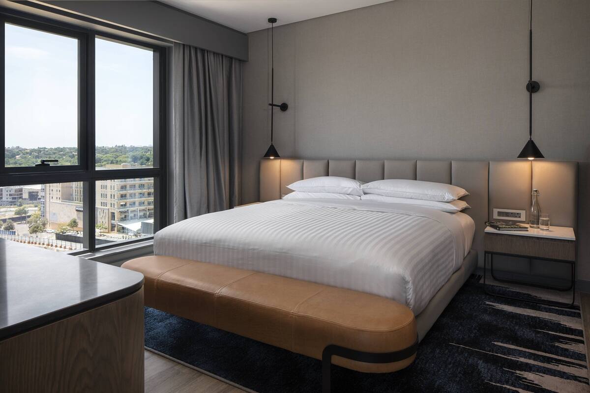 Une chambre d’hôtel moderne dotée d’un grand lit, d’une banquette, d’un bureau et offrant une vue sur la ville grâce à de grandes fenêtres.