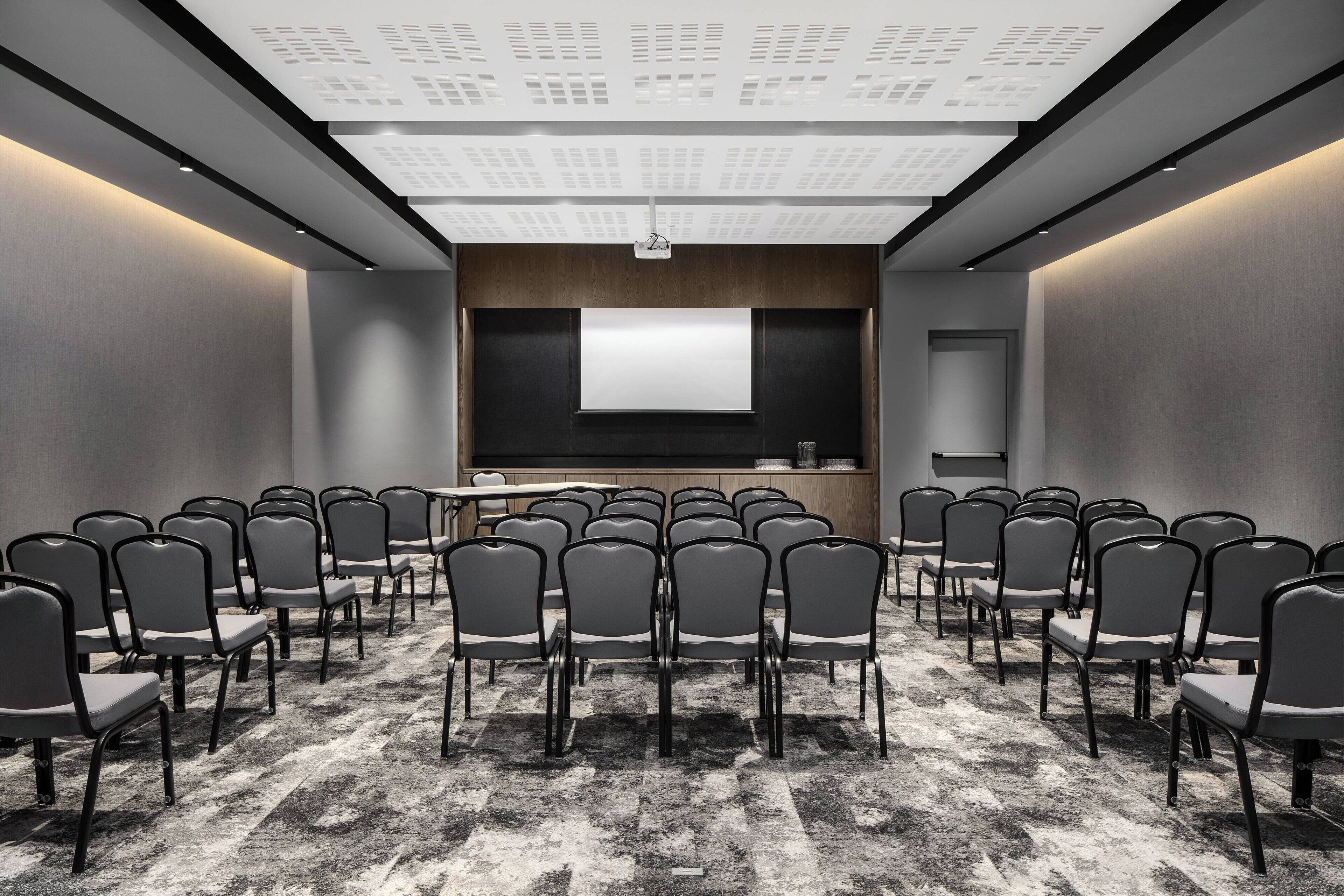 Une salle de conférence avec des rangées de chaises disposées face à un écran de projection, un pupitre et un sol recouvert de moquette.