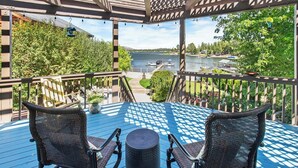 Eine überdachte Terrasse mit zwei Korbstühlen und einem Tisch, mit Blick auf einen See mit Booten und einer anlegenden Pier.