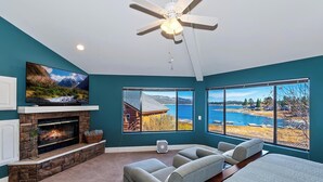Ein Schlafzimmer mit Kamin, Deckenventilator und großen Fenstern mit Blick auf einen See und Berge.