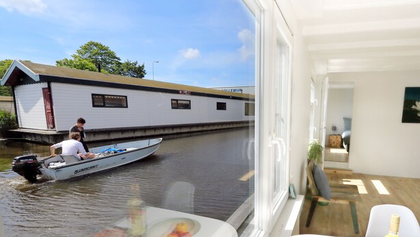 A view from a boat to a white building with a red door, a table with a bowl of fruit, and a bottle on the table.