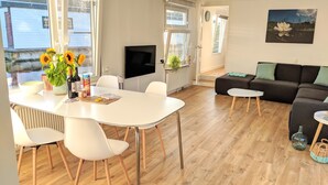 A modern living room with a white dining table, white chairs, a black sofa, and a TV.