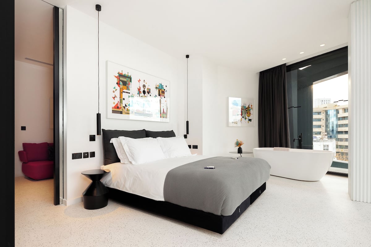 Un dormitorio moderno con una cama grande, una mesita de noche negra, una pared blanca con obras de arte, una bañera negra y vistas a edificios a través de una ventana.