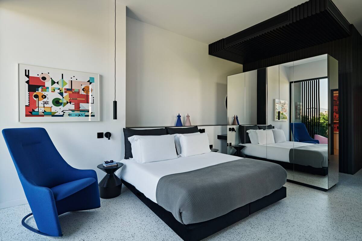 Un dormitorio moderno con una cama grande, un sillón azul, una mesita auxiliar pequeña y un espejo amplio.