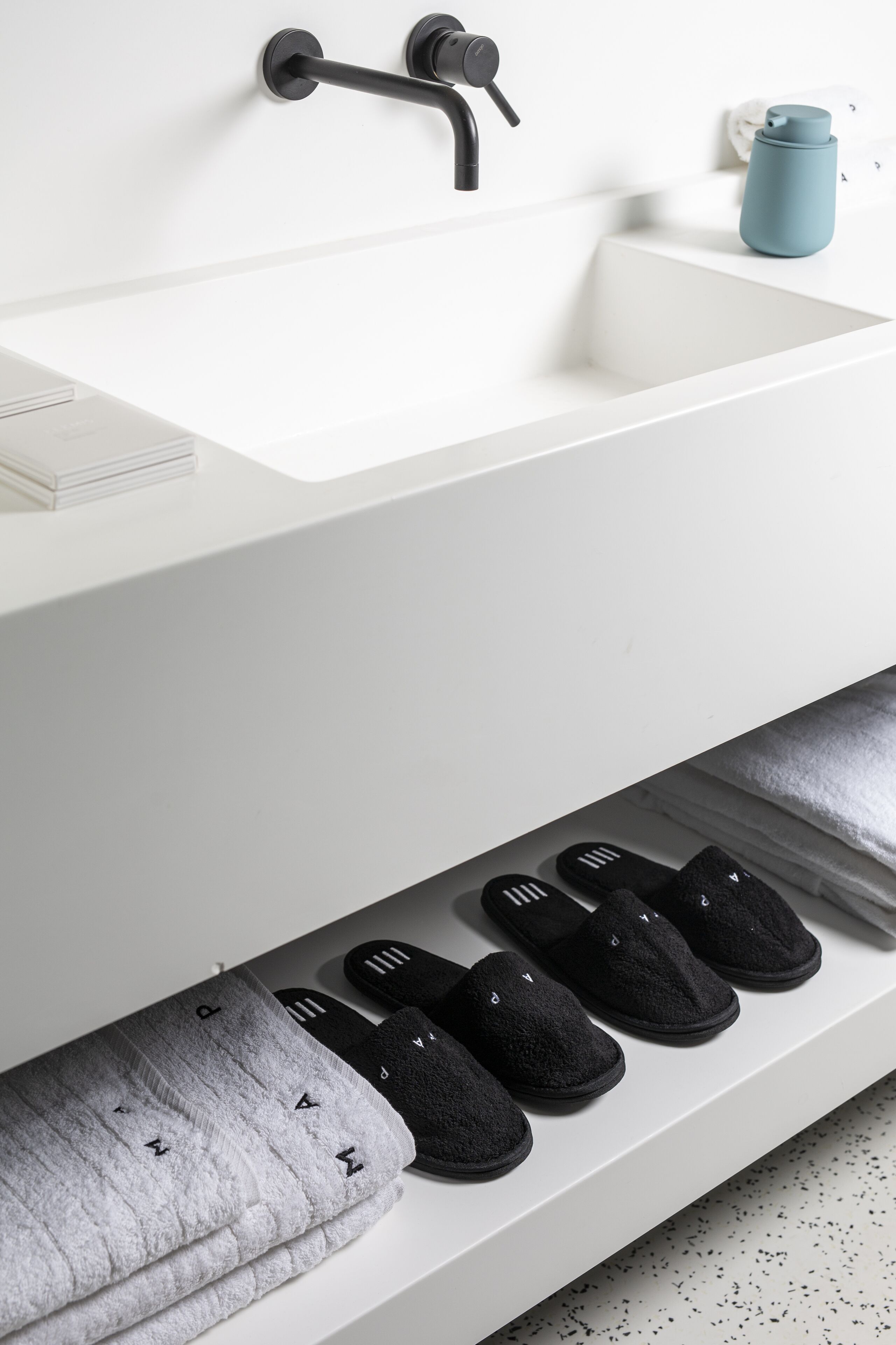 Une salle de bain blanche avec un robinet noir, un lavabo et une étagère sur laquelle reposent des pantoufles noires.