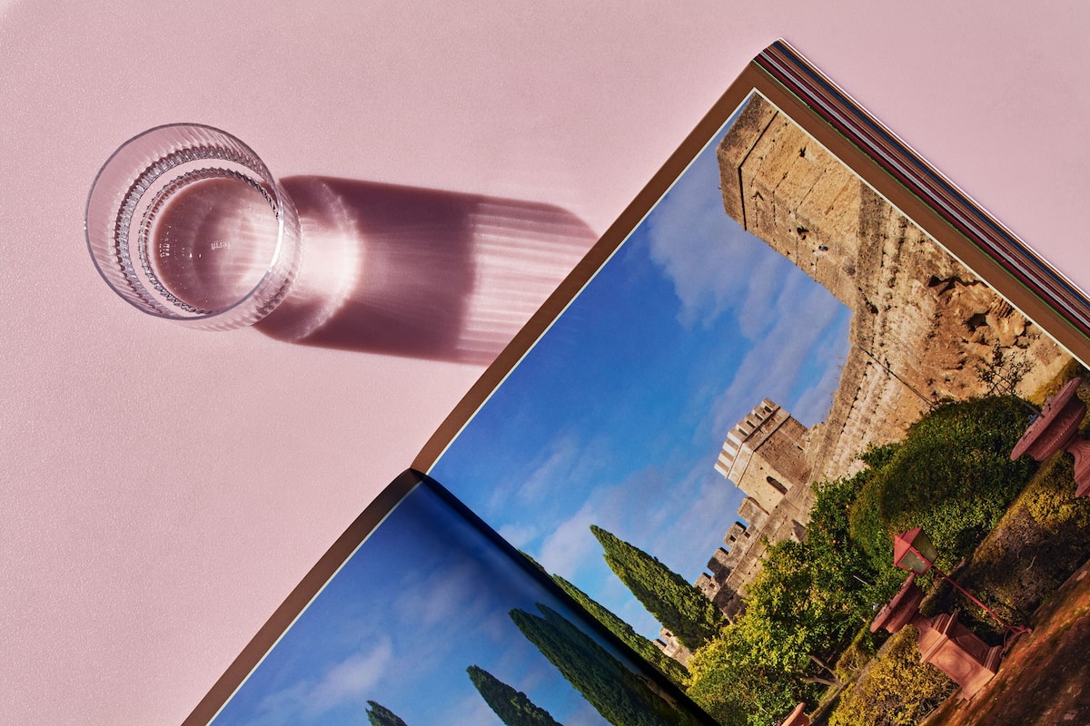 Un vaso de agua sobre una superficie rosa, con una fotografía de un castillo y árboles.