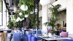 Interior de restaurante moderno con asientos azules, mesas blancas, plantas colgantes y ventanas amplias.