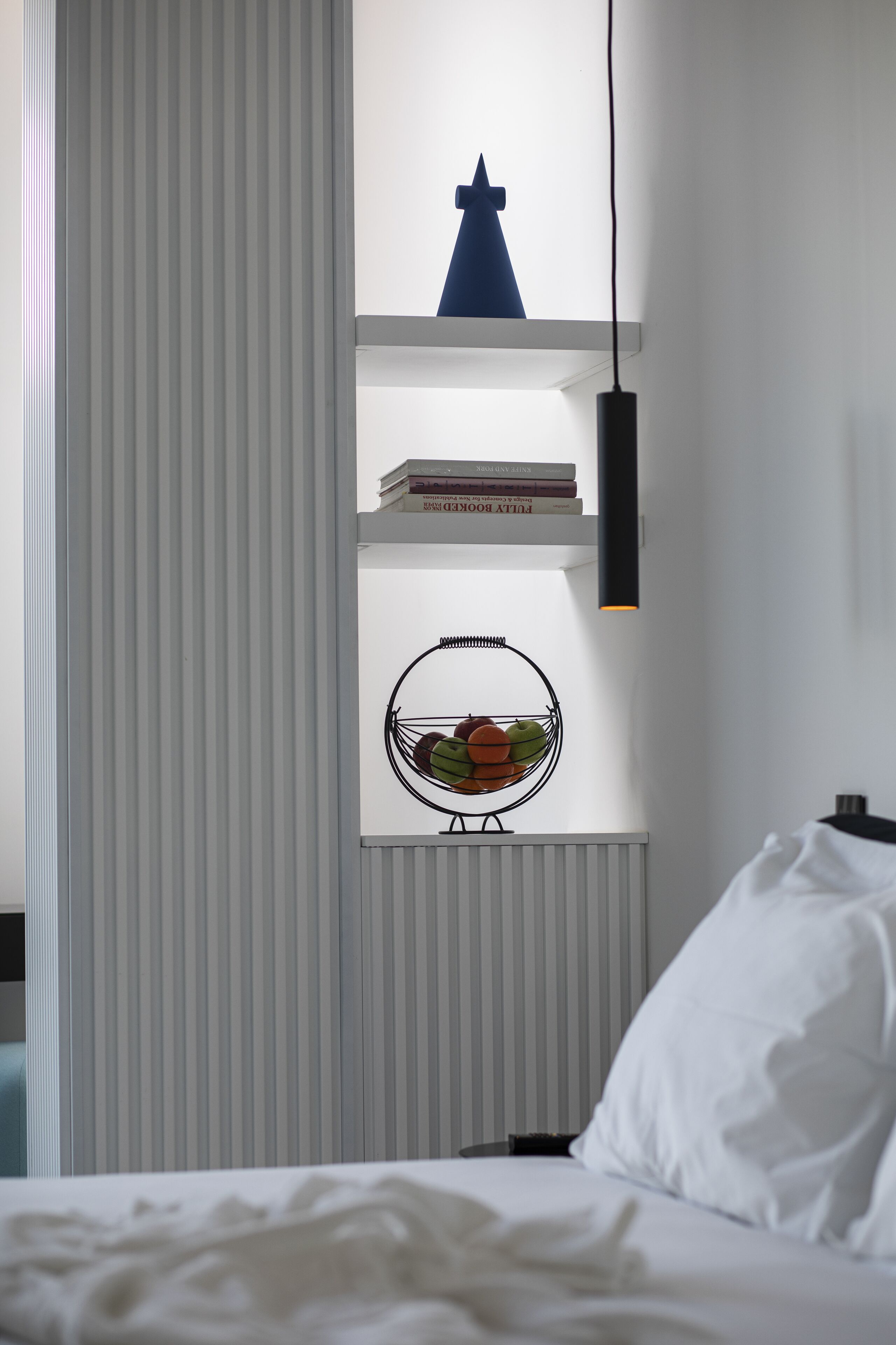 Une chambre à coucher comprenant un lit, une table de chevet, une étagère murale avec des livres et une assiette décorative, ainsi qu’un luminaire suspendu.