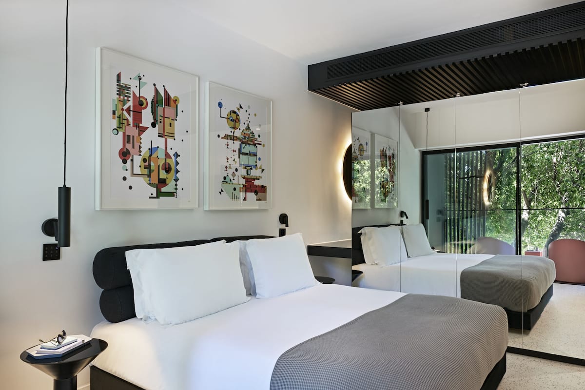 Una habitación de hotel moderna con dos camas, obras de arte en las paredes, un ventanal con vistas a árboles y un ventilador de techo.