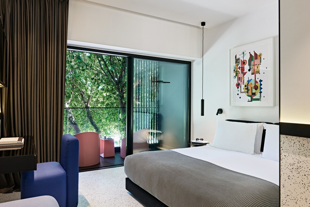 A modern hotel room with a large bed, a desk, a chair, and a balcony with a view of trees.