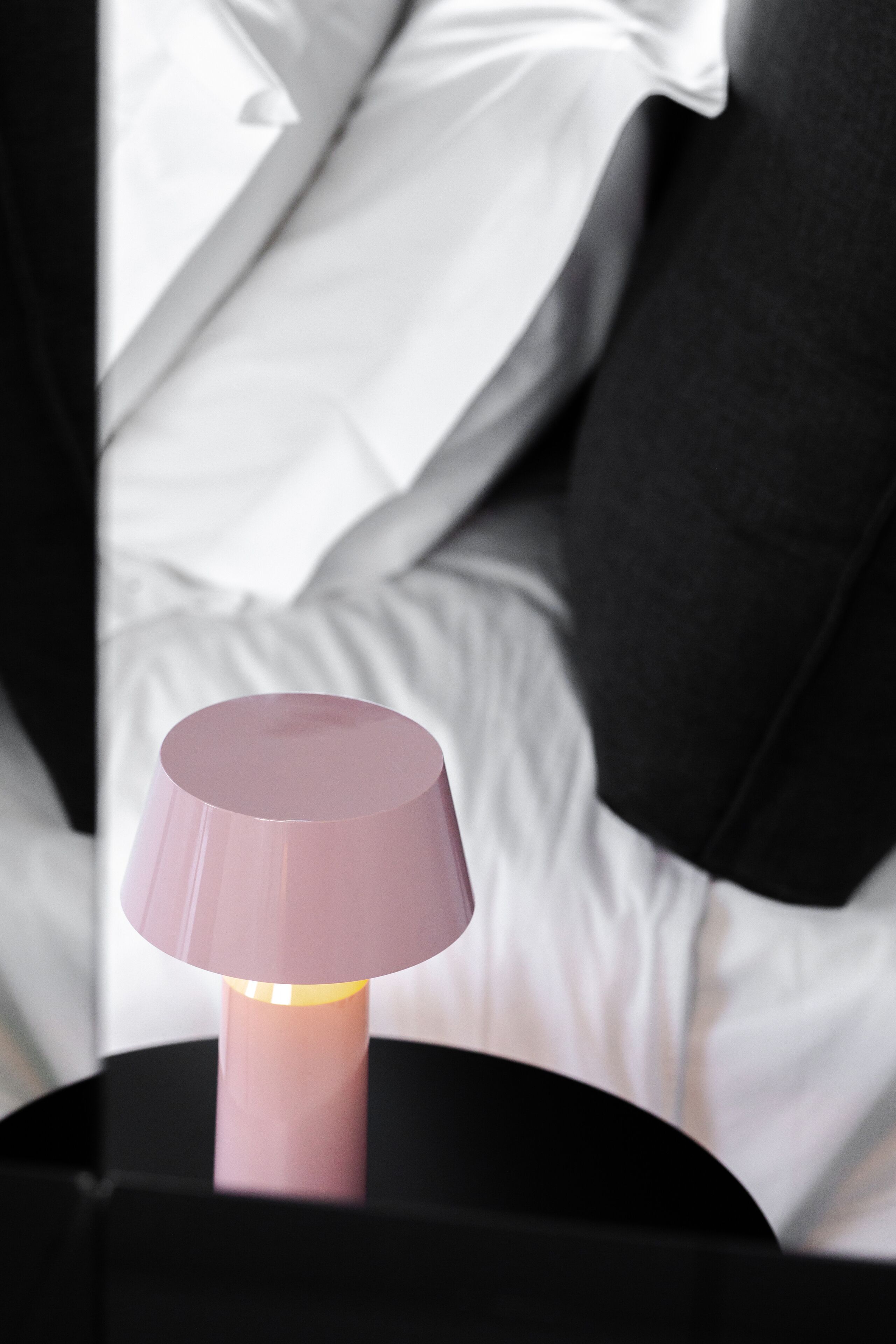 Una cama con ropa de cama blanca y negra, una mesita de noche negra con una lámpara rosa, y un objeto negro sobre la mesita.