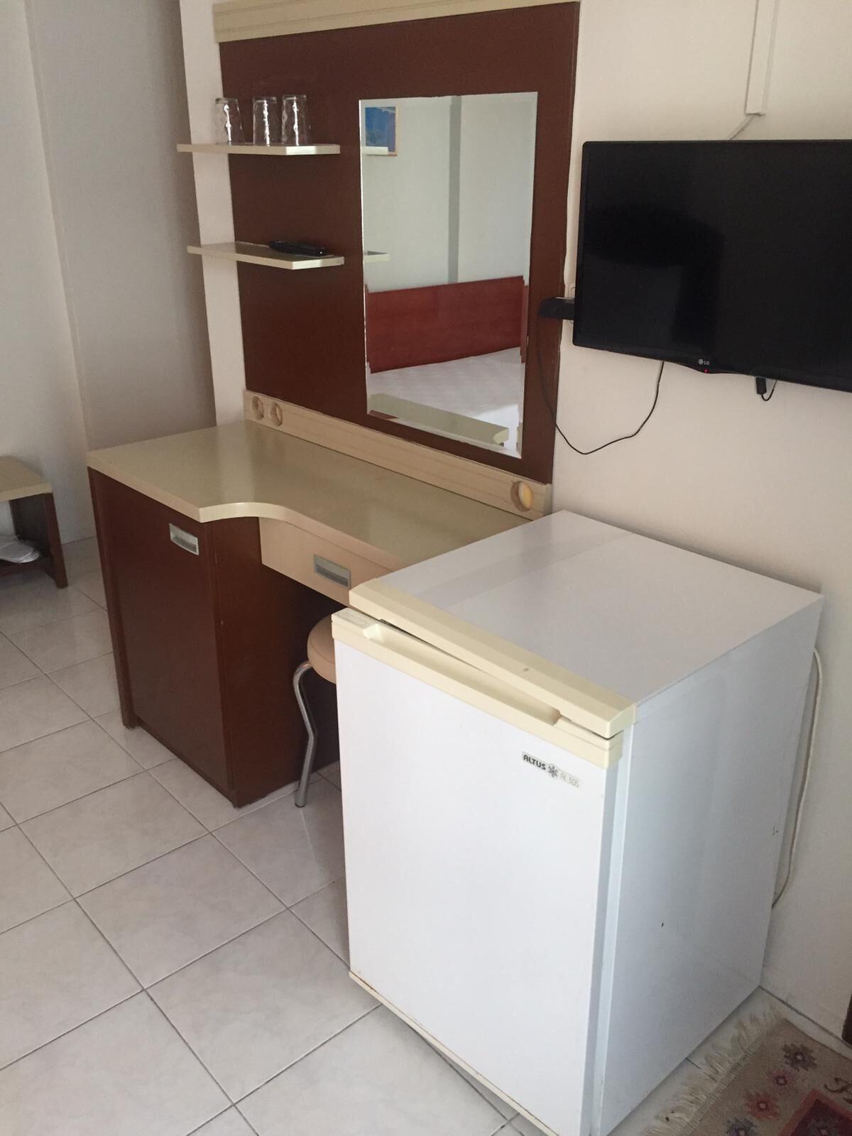 A compact kitchenette with a white refrigerator, a wooden counter, a mirror, and a mounted flat-screen TV.