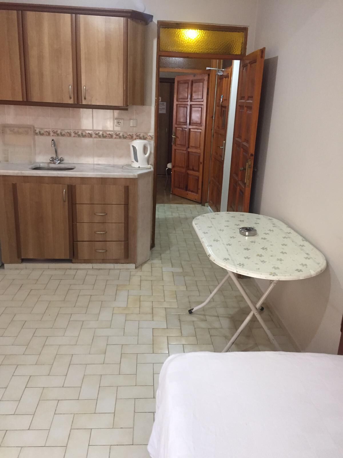 Een gezellige, kleine keuken met houten kastjes, een spoelbak en een waterkoker. Op de voorgrond staat een klein tafeltje met een dienblad en een bed.