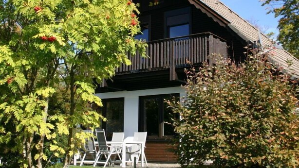 A two-story wooden house with a balcony, surrounded by trees and a lawn.