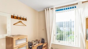 Un dormitorio con escritorio de madera, una silla, una cama, un radiador, una ventana con cortinas, un perchero y un espejo.