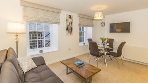 A living room with a sofa, a coffee table, a dining table with chairs, a lamp, and a painting on the wall. - Below Deck by Pureserviced (Plymouth)