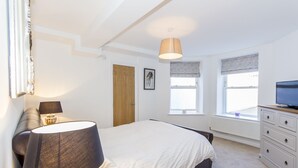 A bedroom with a bed, bedside lamps, a dresser, a television, and a window with blinds. - Below Deck by Pureserviced (Plymouth)