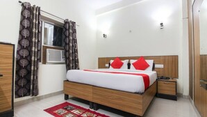 Habitación de hotel con una cama de madera, almohadas rojas, alfombra roja y blanca, una mesita de noche y una ventana con cortinas.