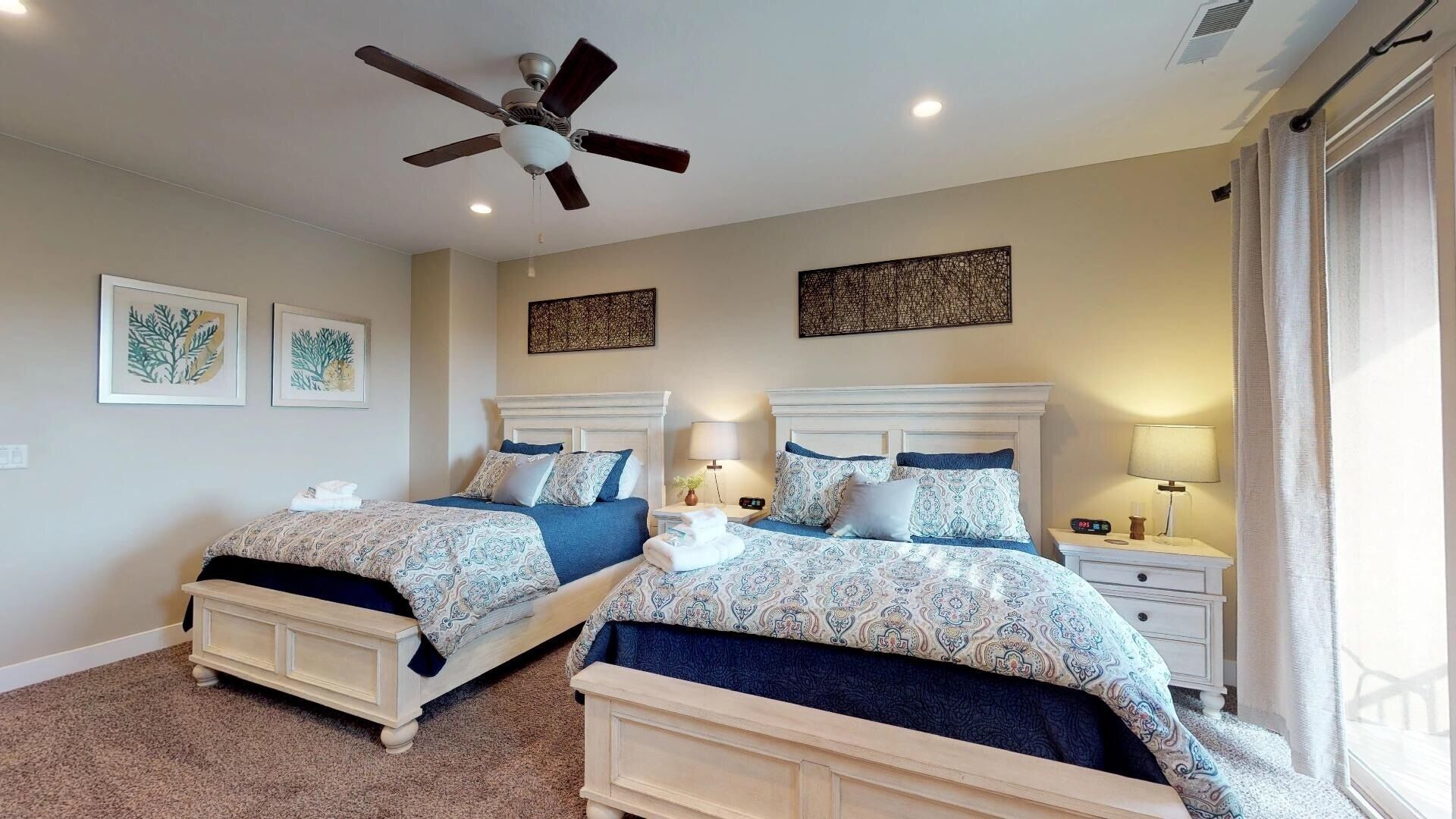 A bedroom with two beds, a ceiling fan, bedside lamps, and artwork on the walls.