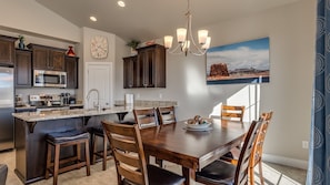 A modern kitchen with dark wood cabinets, a stainless steel microwave, and a granite countertop. There is a dining area with a wooden table and chairs, and a chandelier above.