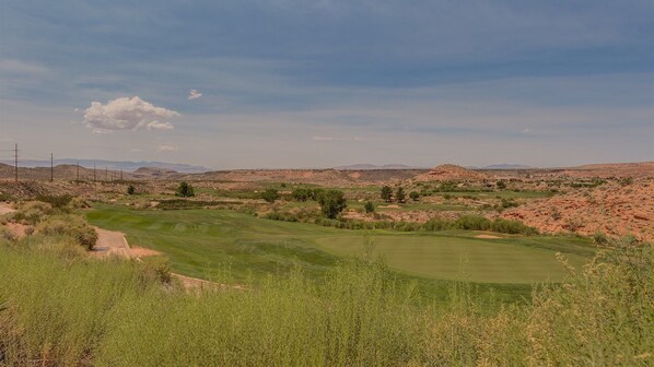 Un campo de golf con cielo despejado, montañas a lo lejos y vegetación escasa.