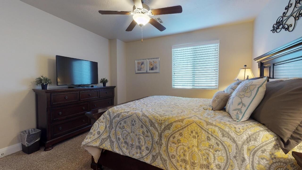 A bedroom with a bed, a dresser, a television, a ceiling fan, and a window with blinds.