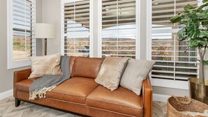 A room with a brown leather sofa, white window blinds, a potted plant, and a lamp.