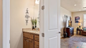 A bathroom with a wooden vanity, a sink, and a mirror. There is a door leading to a living room with a sofa, a coffee table, and a lamp. - Canyon Oasis 4 Bedroom Apts by RedAwning (Washington)