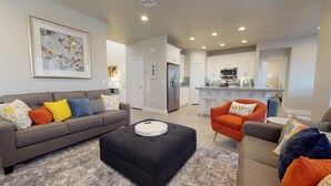 A modern living room with a grey sofa, black ottoman, and orange armchair. A kitchen with white cabinets and stainless steel appliances is visible in the background.