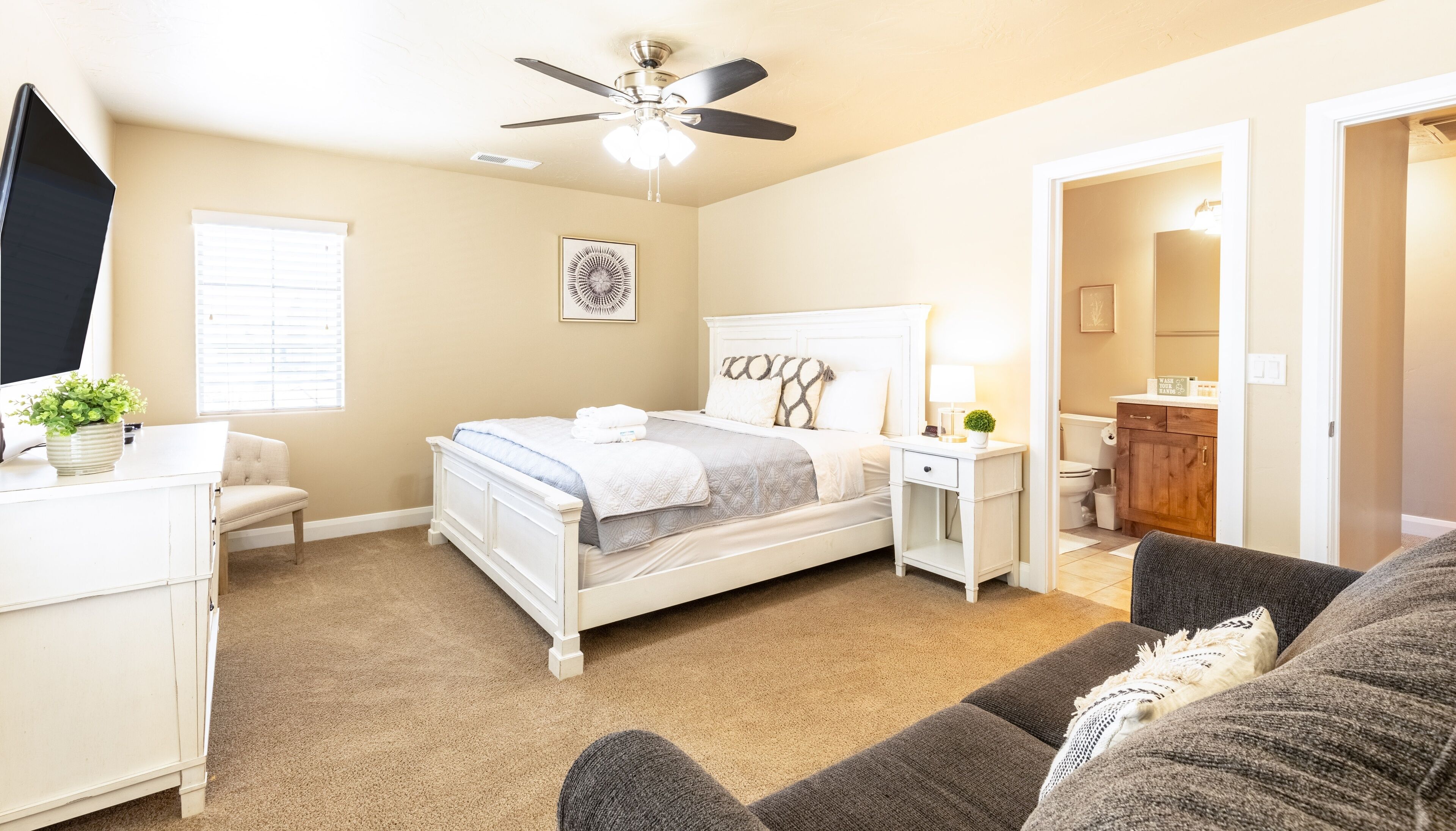 A bedroom with a bed, bedside tables, a ceiling fan, a window with blinds, a framed picture on the wall, and a doorway leading to a bathroom.