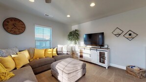 A living room with a sectional sofa, ottoman, television, and a clock on the wall. - Southern Comfort 3 Bedroom Apts by RedAwning (Washington)