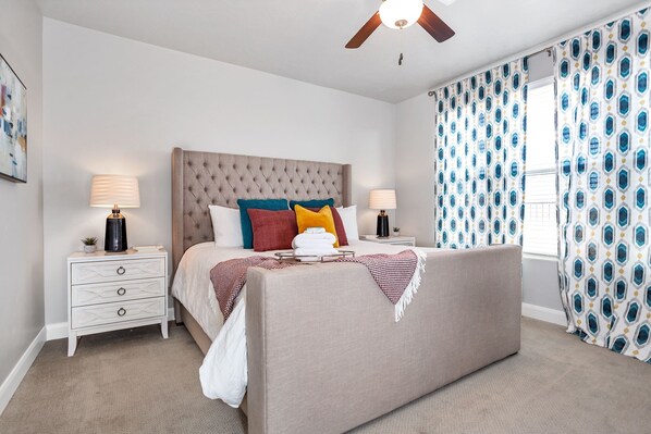A bedroom with a tufted headboard, a bed with multiple pillows, two bedside tables with lamps, a ceiling fan, and patterned curtains. - The Florentine 3 Bedroom Apts by RedAwning (Washington)