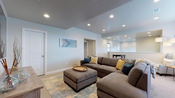 A modern living room with a sectional sofa, ottoman, and a coffee table. There is a dining area with a table and chairs, and a kitchen in the background. - Endless Adventures 4 Bedroom Apts by RedAwning (Washington)